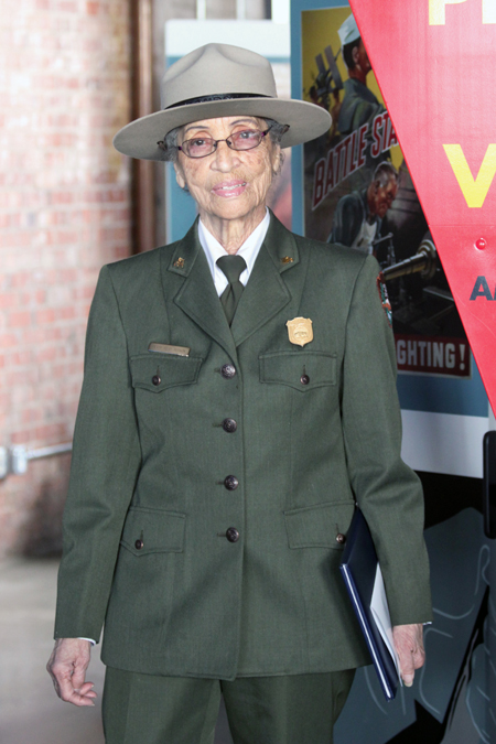 Betty Reid Soskin wearing her park uniform, holding a folder in her hand, and standing in front of posters at the park.